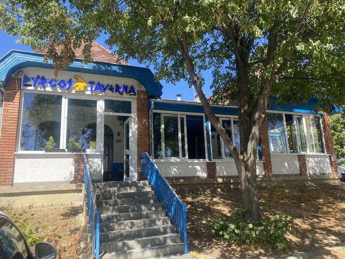 Az étterem bejárata: kék korlátos lépcső vezet fel a PYRGOS TAVERNA cégér alatti ajtóhoz, jobbra az üvegezett veranda