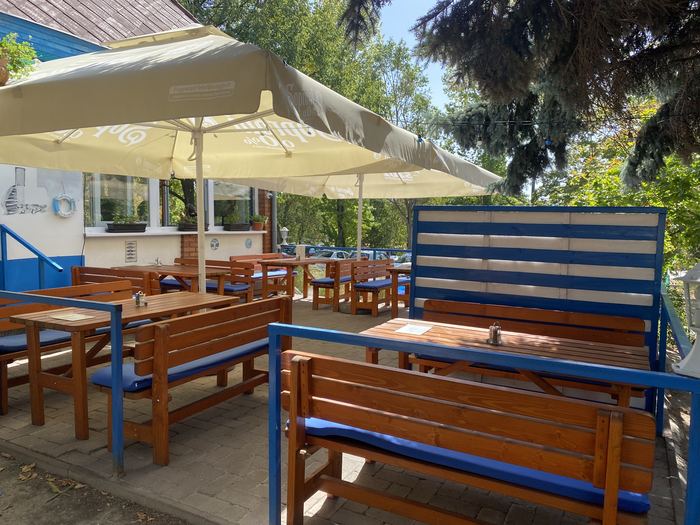 A Pyrgos Taverna terasza: fa asztalok és padok kék üléspárnákkal, krémszínű napernyők alatt, kék-fehér csíkos szélfogó paravánokkal