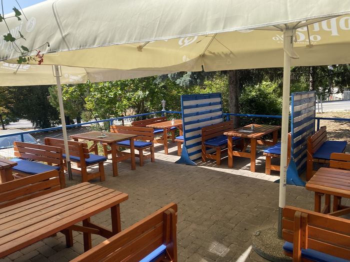 A terasz másik szögből, a napernyők alól: fa asztalsorok kék üléspárnákkal, térköves burkolat, kék korlát és fák