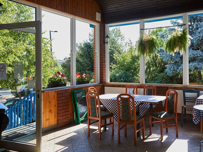 A fedett veranda: faburkolatú falak és mennyezet, körbefutó nagy ablakok a zöldre, függő páfrányok, muskátlik az ablakpárkányon, kockás abroszos asztalok