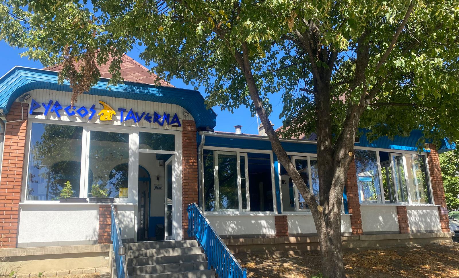 A Pyrgos Taverna épülete Budaörsön, a Szabadság úton: kék-fehér homlokzat, cégér a bejárat fölött, árnyékot adó fákkal